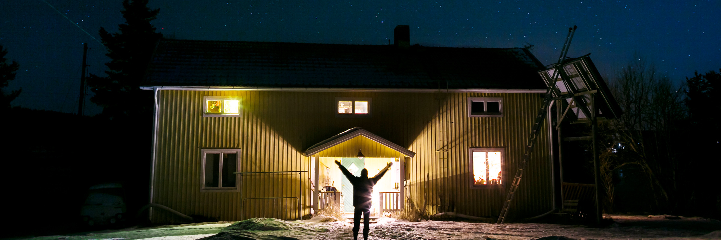 Person sträcker händerna mot himlen framför gult trähus nattetid