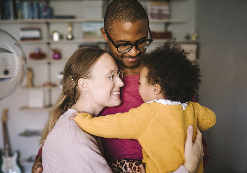 Familj med mamma, pappa och litet barn omfamnar varandra inomhus